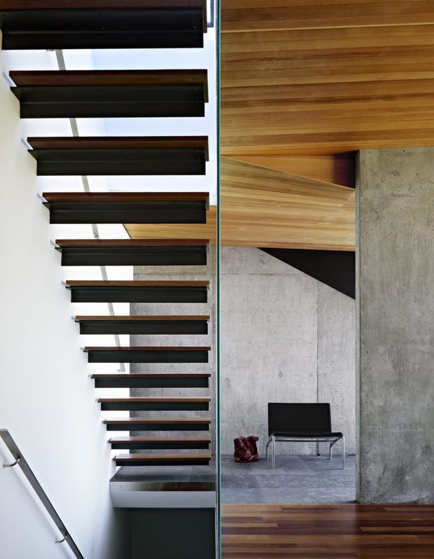 Interior architectural photograph looking along a floating staircase with dark timber treads and slim metal supports beside a glass balustrade. The stair rises alongside white and exposed concrete walls into a warm timber-lined ceiling and landing, with a black chair in the adjacent minimalist space. The composition highlights strong horizontal lines, exposed materials, and carefully detailed contemporary construction.