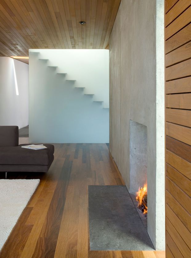 Interior view of a refined contemporary living area with a built-in concrete fireplace, dark stone hearth, warm timber flooring, and a slatted timber ceiling. A minimalist floating stairway is visible behind a frosted glass or pale partition, while a dark sofa and light rug occupy the foreground. Vertical timber wall panelling adds warmth to the right side.