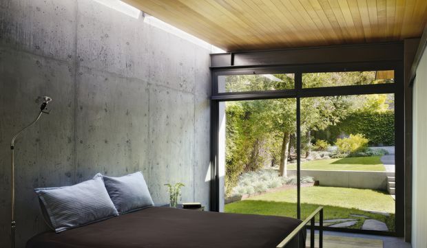 A minimalist bedroom with a low bed positioned against a raw poured-concrete feature wall. Full-height black-framed glazing opens views to a landscaped garden, while a warm timber-lined ceiling adds contrast. The room has a restrained palette of concrete grey, dark bedding, black framing and natural timber, with a small bedside plant and an exterior stair visible beyond the glazing.