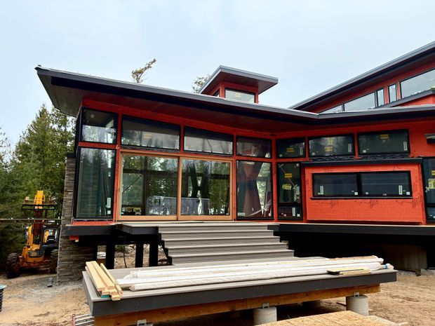 Exterior view of a contemporary timber-framed house under construction, featuring bold red and dark window and door frames, extensive glazing, a low-pitched intersecting roofline, raised deck structure, and broad exterior stairs. Construction materials and equipment are visible in the foreground and beside the building.