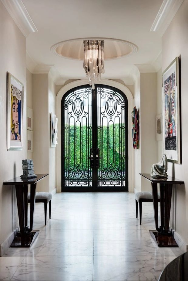 Elegant residential entry hall with a symmetrical layout, polished pale marble flooring, cream walls, ornate white ceiling mouldings, black console tables, contemporary artwork, and a dramatic arched black wrought-iron and glass front door. A suspended crystal chandelier hangs within a circular recessed ceiling detail, creating a refined and gallery-like entrance.