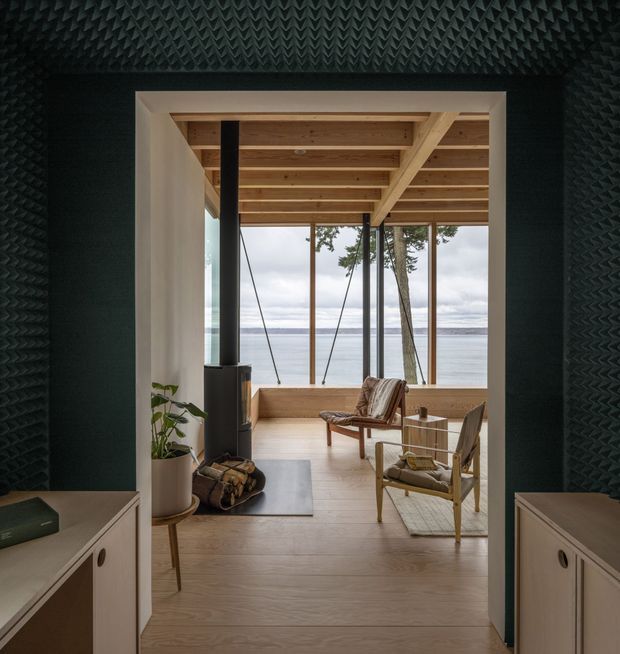 A calm, lake-facing living room viewed through a dark textured entry or hallway. The room features exposed timber ceiling beams, pale timber flooring, full-height glazing with water and trees beyond, a black wood-burning stove, and mid-century-style lounge chairs around a small table. Built-in light cabinetry frames the foreground opening.