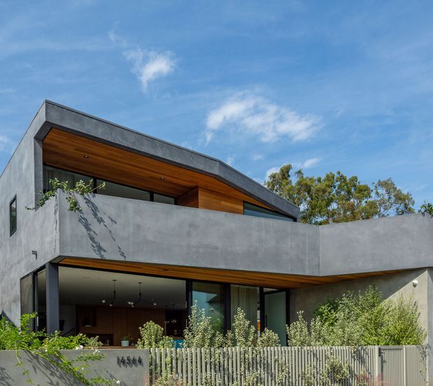 Modern two-storey residential facade with a sculptural flat-roofed form, exposed concrete walls, extensive dark-framed glazing, and warm timber soffits. A projecting upper-level balcony or privacy wall wraps across the front, while landscaped planting and a slatted metal boundary fence define the street edge.