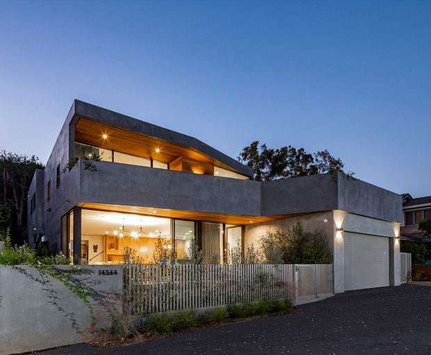 Twilight exterior view of a contemporary two-storey residence with a sculptural flat roof and angular projecting upper volume. The home has smooth grey concrete-like walls, extensive ground-floor glazing, warm timber-lined soffits, a covered entry terrace, vertical metal fencing, and an integrated single garage. Warm interior lighting contrasts with the cool blue evening sky.