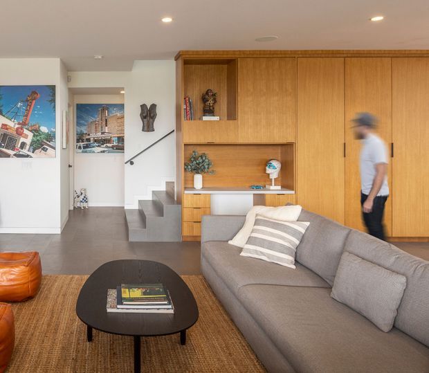 Contemporary living room with a large grey sectional sofa, rounded black coffee table, and warm timber built-in cabinetry spanning the rear wall. The joinery includes open display niches, shelving, and concealed storage. A short set of concrete or grey tiled steps leads toward a hallway, with framed artwork and sculptural decor adding visual interest. Recessed ceiling lights provide even illumination, while natural timber, neutral upholstery, and textured flooring create a warm, refined atmosphere.
