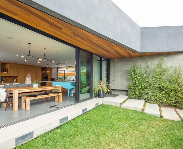 View from a landscaped courtyard into a contemporary open-plan dining and living area through fully retractable dark-framed glazing. The home features a flat-roofed concrete form, warm timber soffit and interior wall panelling, polished concrete-like flooring, and a minimalist courtyard with lawn, concrete stepping slabs, and planting. A timber dining table and benches, kitchen joinery, and colourful poolside furniture are visible inside.