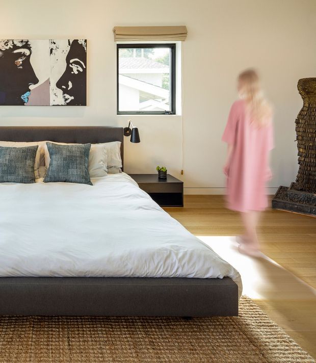 A bright, contemporary bedroom with a low-profile upholstered bed, white bedding, textured blue-grey cushions, and a dark bedside table with a black reading lamp. A black-framed window with a timber Roman blind brings in natural light, while a large monochrome artwork and woven rug add visual texture. A sculptural woven chair or screen sits in the corner, and a blurred person is walking through the room.