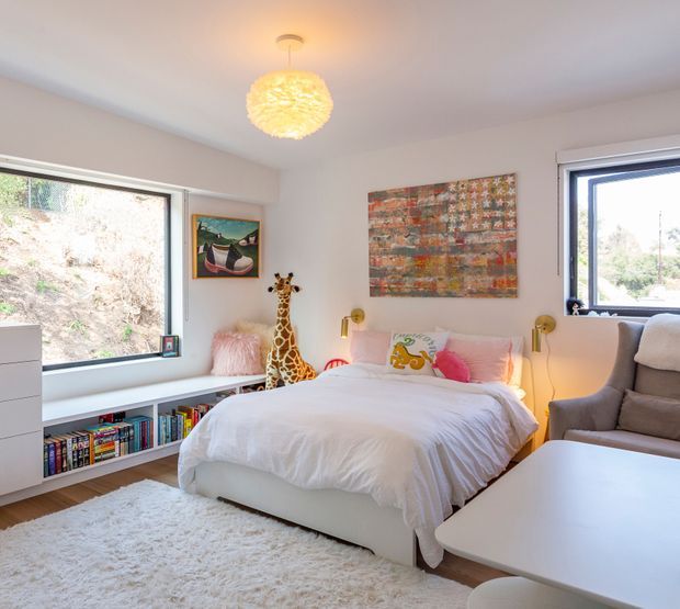 Bright, cozy bedroom with a white upholstered bed, built-in low window-seat shelving filled with books, framed artwork, a plush rug, a grey armchair, and a sculptural pendant light. Large windows provide natural light and overlook a wooded hillside. Playful decorative accents include a giraffe figure and colourful cushions.