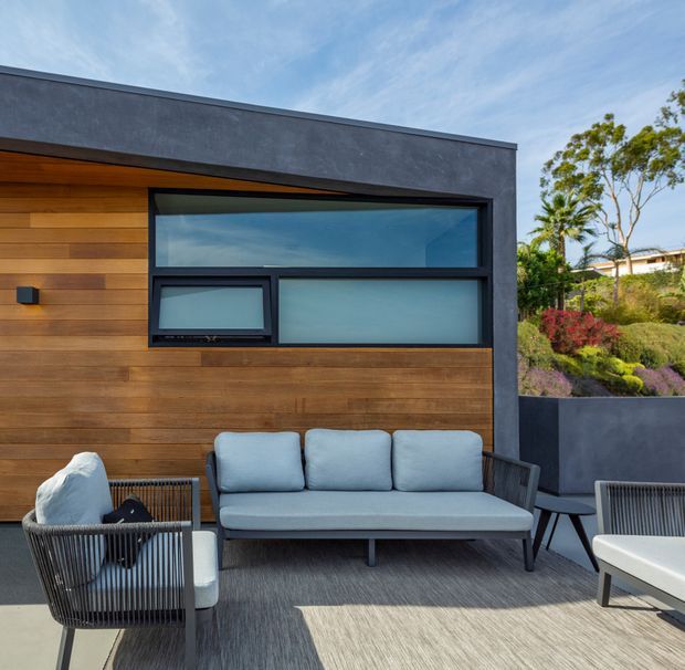 A contemporary outdoor lounge terrace beside a modern single-storey structure. The facade combines dark grey rendered wall sections with horizontal warm timber cladding, black-framed glazing, and a broad roofline. A pale blue-grey sofa and matching lounge chairs sit on a light-toned deck or paved terrace, with landscaped planting visible beyond.