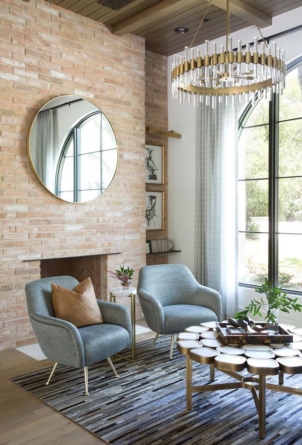 A refined living room with an exposed pale brick feature wall, integrated fireplace, round wall mirror, tall black-framed arched glazing, and timber slatted ceiling. Two textured blue-grey lounge chairs sit on a striped area rug around a sculptural multi-tier coffee table, while a large brass-and-glass chandelier provides a decorative focal point. The room has abundant natural light and a warm, sophisticated material palette.
