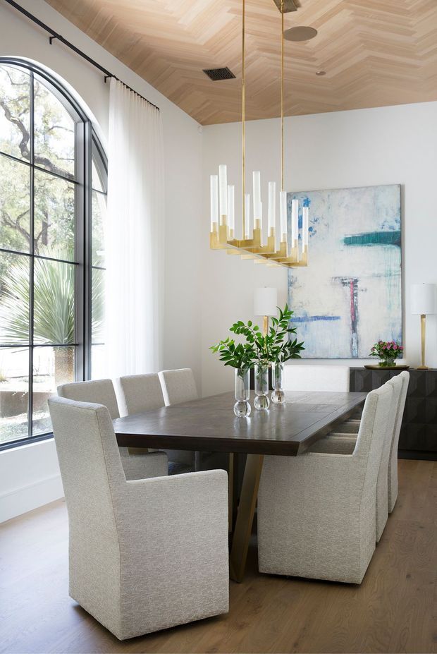 A bright, refined dining room with a large dark timber dining table surrounded by upholstered cream dining chairs. The room features a dramatic arched black-framed window with sheer curtains, white walls, a warm herringbone timber ceiling, pale timber flooring, a sculptural brass and glass pendant light, contemporary abstract artwork, and a dark built-in sideboard. Greenery and natural light add an organic touch.