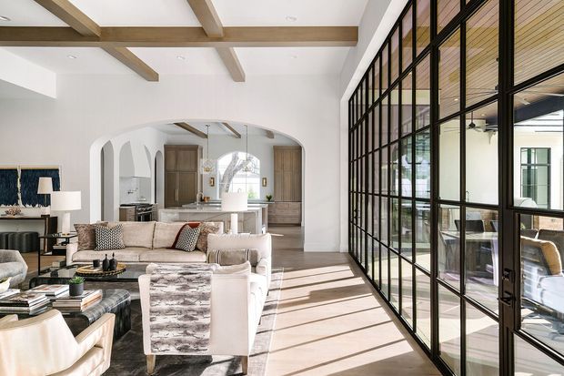 Bright open-plan living room with a large seating area, exposed timber ceiling beams, white walls, and a kitchen visible through a broad arched opening. A full-height black-framed glazed partition and door line one side of the room, revealing an adjacent dining or workspace area. The space has layered neutral furnishings, warm timber details, and strong natural light creating a refined contemporary interior.