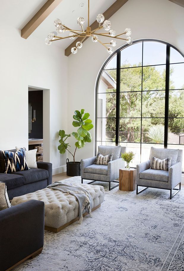 A bright, spacious living room with a very high vaulted ceiling, exposed timber beams, and a dramatic black-framed arched window with extensive glazing. The room contains a dark upholstered sofa, two light grey armchairs, a tufted cream ottoman, patterned cushions, a large pale patterned rug, a potted fiddle-leaf fig, and a sculptural brass-and-glass chandelier. A dark built-in area is visible through a doorway at left.