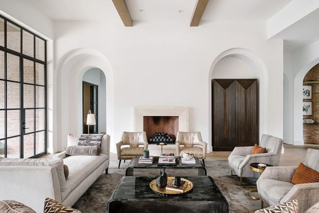 A refined, spacious living room with white plaster walls, tall arched openings, exposed timber ceiling beams, large black-framed steel-look glazing, and a dark carved timber door. The room is furnished with light upholstered sofas and armchairs around layered dark stone or glass coffee tables, with a central fireplace and neutral patterned rug. The overall scheme is warm, elegant, and contemporary with traditional architectural detailing.