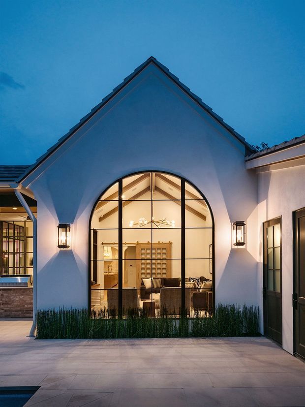 Dusk exterior view of a refined white stucco residence centered on a steep gabled facade. A large black-framed arched glass opening reveals a warmly lit living room and exposed timber ceiling structure inside. Matching wall lanterns flank the opening, with dark-framed glazed doors to the right, landscaped grasses at the base, and pale paved flooring in the foreground. An adjoining glazed wing and partial pool area are visible to the left.