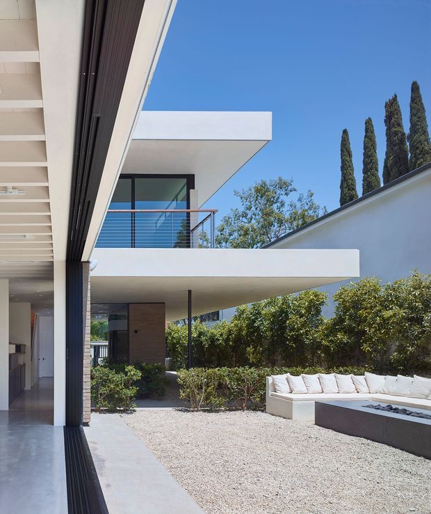 A bright contemporary courtyard view of a minimalist multi-level residence. White cantilevered concrete slabs and flat roof forms frame dark-framed glazing, a timber-toned entry wall, and slim metal balcony railings. The gravel courtyard contains built-in outdoor seating and a long rectangular fire feature, with tall narrow trees and clipped hedges around the perimeter. A covered indoor-outdoor threshold with dark vertical screening is visible at left.