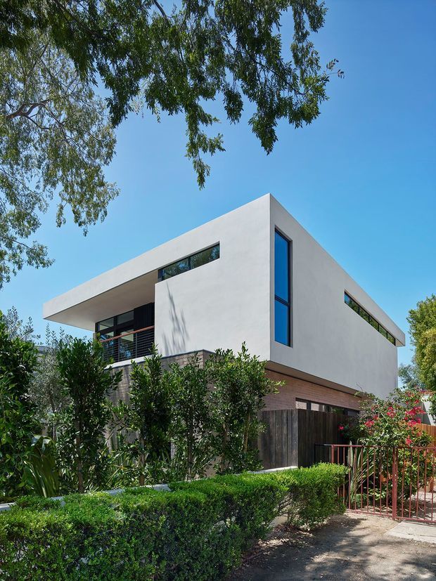 Exterior view of a contemporary two-storey residence with a crisp white cubic upper volume, narrow horizontal strip windows, a tall vertical window, dark glazing, and a projecting flat roofline. The lower level combines warm brickwork with dark timber screening and fencing. Dense hedges, mature trees, and a red metal pedestrian gate frame the landscaped front approach.