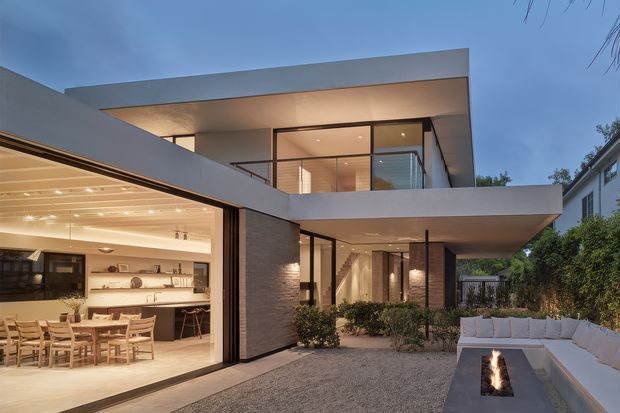 Contemporary two-storey residence viewed from a landscaped courtyard at dusk. The home features strong horizontal flat roof planes, extensive floor-to-ceiling glazing, warm interior lighting, dark-framed windows, and textured masonry walls. An open-plan kitchen and dining area is visible through the large glazed opening, while the courtyard includes built-in outdoor seating and a linear fire feature.