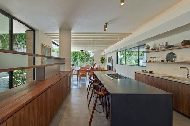 A spacious open-plan kitchen and dining area with a long central island, integrated cooktop, timber cabinetry, and dark stone or solid-surface island base. Floor-to-ceiling glazing and a glazed partition bring in abundant greenery and natural light. Open timber display shelving, warm wood joinery, concrete-look flooring, and a slatted ceiling create a refined contemporary interior.