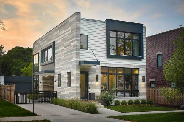 Exterior view of a contemporary two-storey urban residence with a boxy form, flat roof, large dark-framed windows, and a contrasting facade combining light horizontal cladding with pale masonry. The house sits on a landscaped suburban lot with a concrete driveway, front path, fencing, and low planting.
