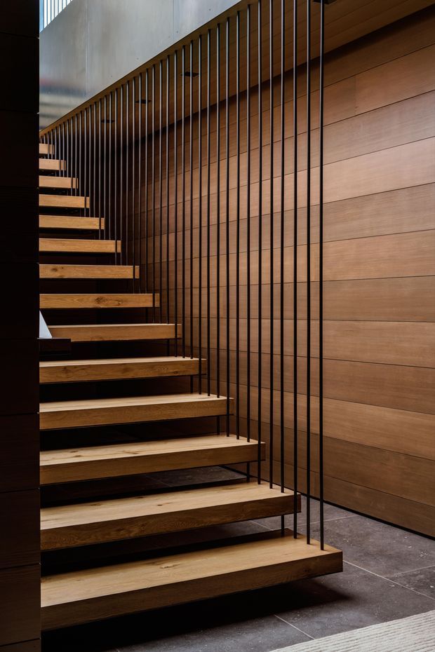 Interior architectural photograph focused on a contemporary stairway. Floating timber treads rise beside a warm horizontal timber-clad wall, enclosed by a slender black vertical metal balustrade. Dark stone or concrete-look floor tiles and a contrasting light ceiling panel complete the refined, minimalist composition.