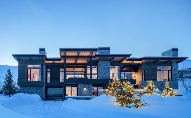 A large contemporary multi-level residence set in a snowy landscape at dusk. The house has a strongly horizontal composition, dark exterior cladding, extensive glazing, projecting flat roof planes, warm timber soffits, exposed stone-clad chimney or wall elements, and an integrated garage. Snow-covered ground and illuminated interiors create a striking contrast with the cool blue exterior light.