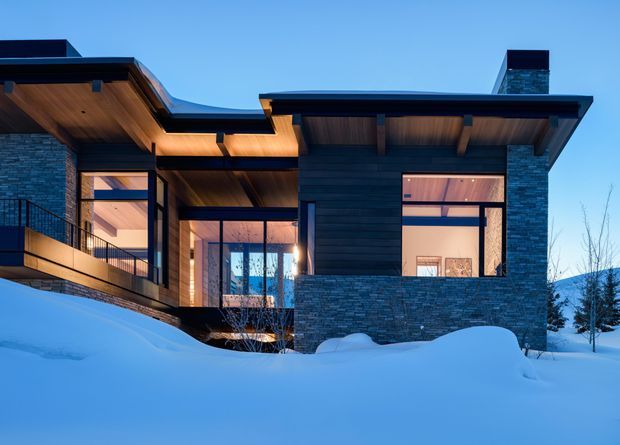 Exterior view of a contemporary multi-level house in a snowy setting at dusk. The home features dark horizontal cladding, extensive stacked-stone masonry, broad overhanging flat or low-pitched roof planes, exposed timber soffits and beams, and large illuminated windows. Deep snow covers the foreground and surrounds the building.