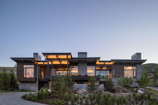 Wide exterior view of a contemporary multi-level residence set into a landscaped site. The house features strong horizontal roof planes, extensive glazing, dark timber-toned cladding, stacked grey stone wall sections, projecting balconies, and warm interior lighting visible through the windows. Native-style planting and boulders occupy the foreground, with low hills in the background.