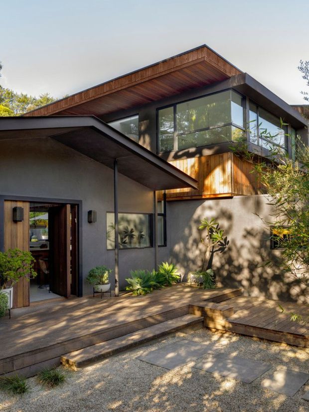 Exterior view of a contemporary two-storey residence with intersecting low-pitched roof planes, deep timber-lined eaves, dark-framed glazing, timber wall cladding, and rendered walls. The entry opens onto a timber deck and stepped gravel courtyard with built-in planting and tropical-style landscaping.