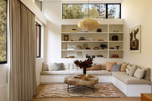 Bright, refined living room with a large L-shaped built-in sectional in warm off-white upholstery, extensive open shelving, tall windows, full-height beige curtains, a sculptural woven pendant light, and a patterned area rug. The room has a clean contemporary shell with warm natural accents and curated display objects.
