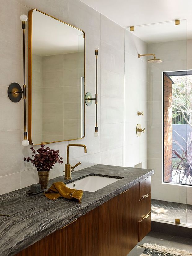 A refined contemporary bathroom with a floating dark timber vanity, dark veined stone benchtop and integrated white basin. Large pale wall tiles continue into the walk-in shower, which has brushed brass fixtures and a frameless glass screen. A rounded-rectangle brass-framed mirror and matching globe sconces add a warm decorative detail, while a tall window brings in views of planted outdoors.