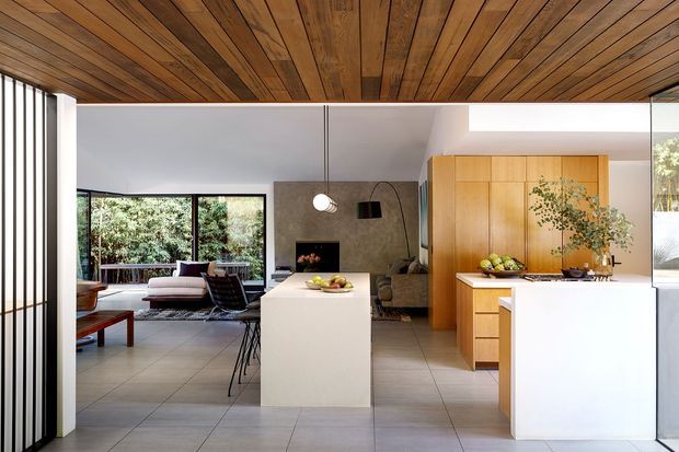 Open-plan contemporary kitchen and living area viewed from the entry, featuring a large white island, warm timber cabinetry, a timber-lined ceiling, grey tiled flooring, expansive glazing, and a compact lounge with fireplace beyond.