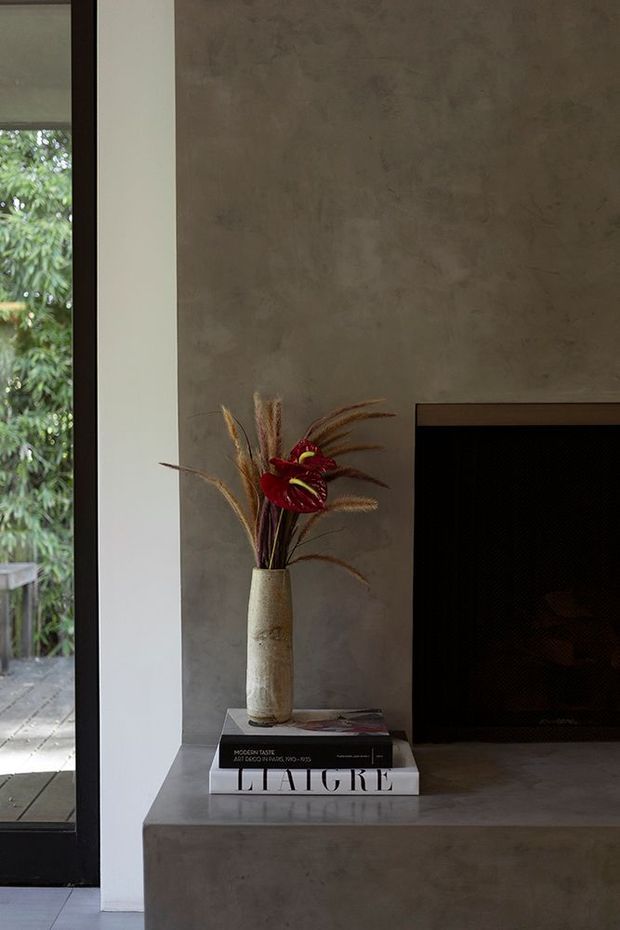A close-up architectural interior detail showing a minimalist concrete fireplace surround and hearth beside a tall dark-framed glazed opening. A textured neutral grey-brown wall is paired with a sculptural ceramic vase and dried botanical arrangement resting on stacked design books. The composition is moody, restrained, and contemporary, with greenery visible outside through the glazing.