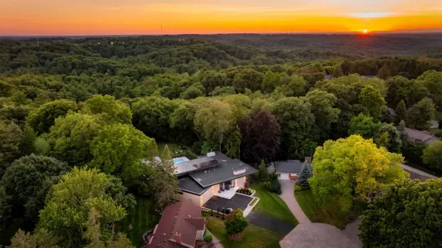 Aerial sunset view of a residential property surrounded by dense mature woodland. The image shows a contemporary flat-roofed main house with extensive glazing and dark roof surfaces, alongside a smaller pitched-roof structure, landscaped lawns, driveways, and a partially visible swimming pool.