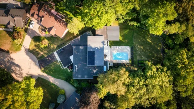 Aerial view of a large residential property surrounded by mature trees and landscaped grounds. The central home has several interconnected roof sections, expansive dark and light roof surfaces, and a bright rectangular swimming pool beside the house. A curved driveway and neighbouring residential structures are also visible.