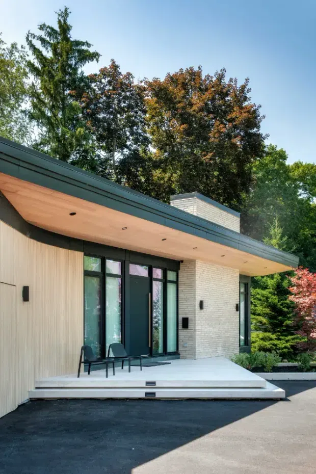 Modern single-storey residential entrance viewed from the exterior, featuring a broad flat roof with a deep timber-lined soffit, dark metal fascia and window frames, pale vertical wall cladding, and a light white-brick feature wall. A raised concrete entry platform leads to a dark front door with sidelights, while mature landscaping surrounds the facade.