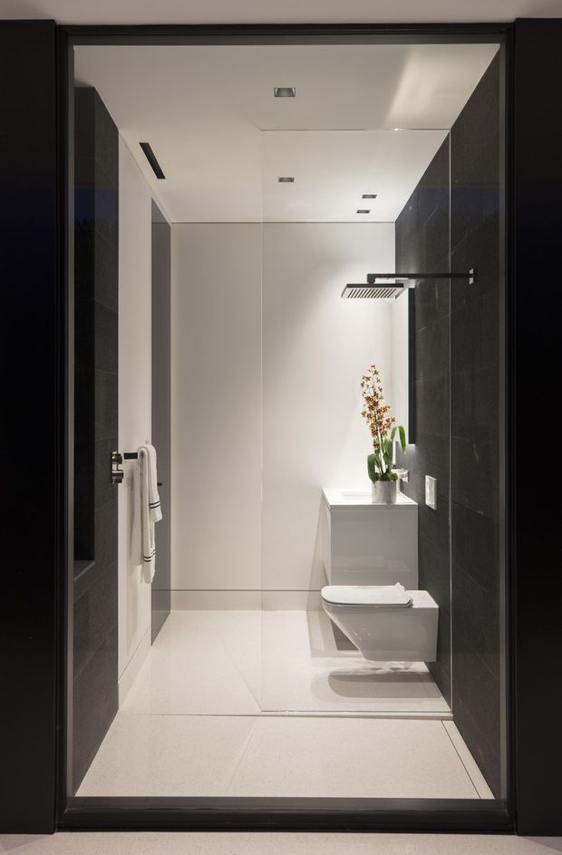 A compact, contemporary bathroom viewed through a dark-framed doorway. The room features a frameless glass shower enclosure with a wall-mounted black rain shower, a wall-hung white toilet, a minimalist white pedestal or concealed cistern enclosure with a potted orchid, large pale floor tiles, dark charcoal wall tiles, white walls and recessed ceiling lighting.