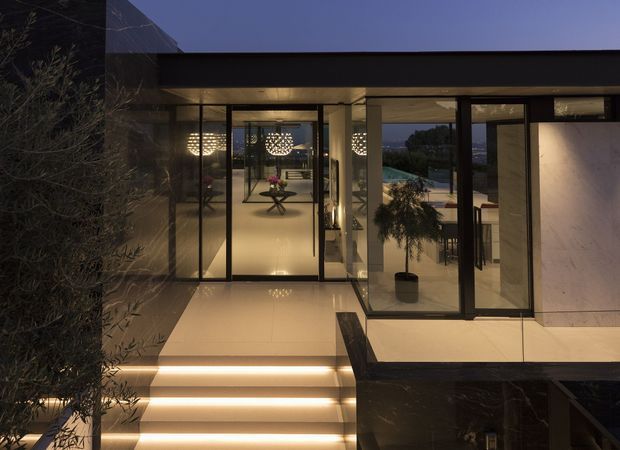 Twilight exterior view of a sleek, multi-level contemporary residence with extensive floor-to-ceiling glazing, dark framed doors and windows, pale stone or tile surfaces, and integrated linear lighting along the entry stairs. The glazed interior reveals a refined living or lounge area with sculptural pendant lighting and a small table, while landscaped planting and a distant residential setting are visible beyond.