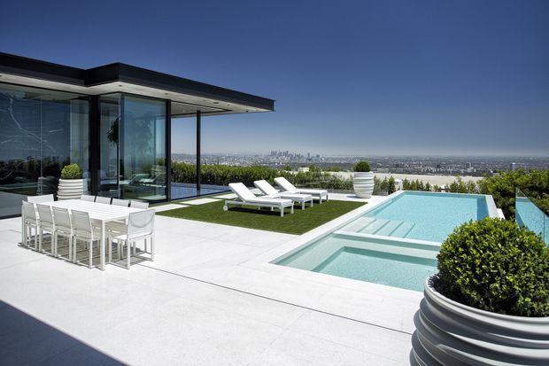 A contemporary rooftop or elevated terrace with a flat-roofed glass pavilion, expansive white-paved outdoor entertaining area, dining setting, sun loungers, and a tiered swimming pool. The property overlooks a broad urban skyline, with minimalist landscaping in large sculptural planters and a clear blue sky.