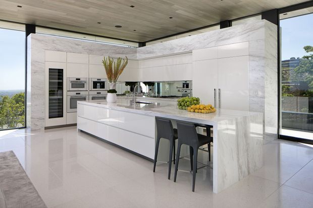 A bright, spacious contemporary kitchen with a large white waterfall island, integrated cabinetry and appliances, a marble-look full-height wall finish, and seating for three at the island. Floor-to-ceiling glazing opens the room to expansive outdoor views. The space has a refined, minimalist aesthetic with light timber ceiling lining and polished pale flooring.
