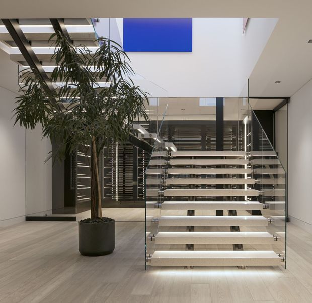 A spacious contemporary interior centered on a floating open-riser staircase with illuminated pale stone or concrete treads and frameless glass balustrades. The stairway rises through a bright double-height void beneath a large skylight, with dark horizontal slatted screens and structural framing behind it. Pale timber-look flooring, white walls, recessed lighting, and a tall potted indoor tree create a refined, minimalist atmosphere.