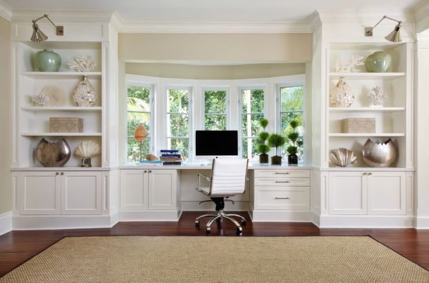 Bright, symmetrical home office with a built-in desk positioned in a bay of windows, flanked by white built-in shelving and lower cabinetry. The room features white painted joinery, decorative crown moulding, dark timber flooring, a large natural woven rug, brass-toned adjustable wall lights, and restrained decorative objects. Garden greenery is visible through the windows.