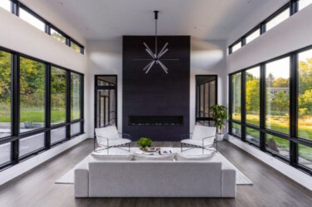 Bright, double-height contemporary living room with extensive floor-to-ceiling black-framed glazing on both sides, a dark feature wall with a linear fireplace and starburst wall clock, and a minimalist seating arrangement facing the fireplace. The room opens visually to a landscaped outdoor setting through the glazing.