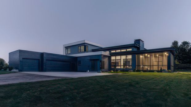 A dusk exterior photograph of a contemporary multi-level residence with a low, angular roofline and dark horizontal cladding. The home features multiple integrated garage doors, large floor-to-ceiling glazed sections, warm interior lighting, and a broad lawn and driveway in the foreground.