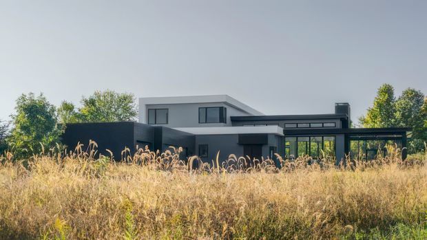 A low, modern flat-roofed house viewed from across a field of tall grasses. The building has a restrained dark and light grey facade, strong horizontal lines, large areas of glazing, and a projecting covered terrace or carport-like structure. Mature trees surround the property, creating a semi-rural setting.