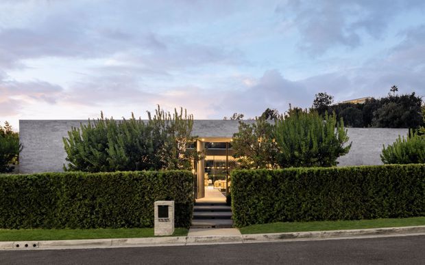 Street-facing view of a contemporary, low-profile residence largely screened by tall manicured hedges. A central glazed entry pavilion with warm interior lighting is reached by a short flight of steps, while broad pale grey rendered walls and a flat roofline are visible behind the landscaping.