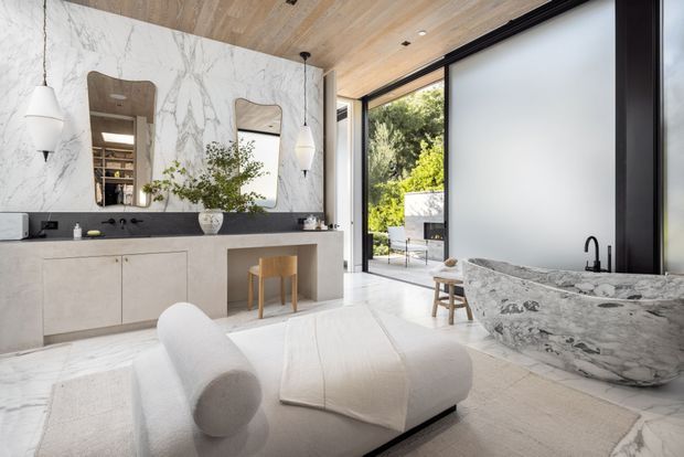 Luxurious contemporary bathroom with a dramatic veined marble feature wall, matching sculptural freestanding stone bathtub, long dark stone vanity, pale integrated cabinetry, and twin wall-mounted pendant lights. A white upholstered lounge chair sits on a neutral rug in the foreground. Timber flooring and ceiling add warmth, while full-height glazed doors open to a landscaped outdoor area.