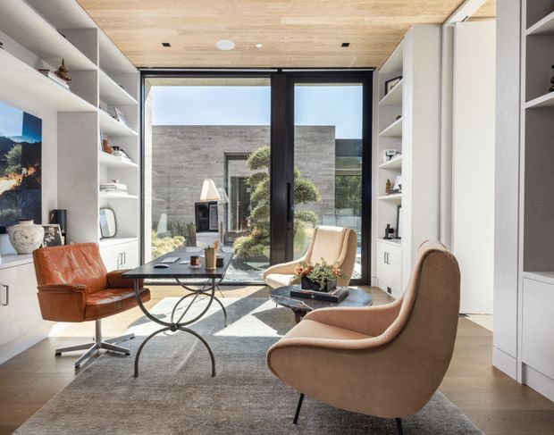 A refined modern living room or lounge with floor-to-ceiling black-framed glazing opening to a landscaped courtyard. Built-in white shelving and cabinetry line both sides of the room, with a warm timber ceiling, pale wood flooring, a large neutral rug, sculptural coffee table, and two tan leather or upholstered lounge chairs. The space is bright, carefully detailed, and visually connected to the exterior courtyard.