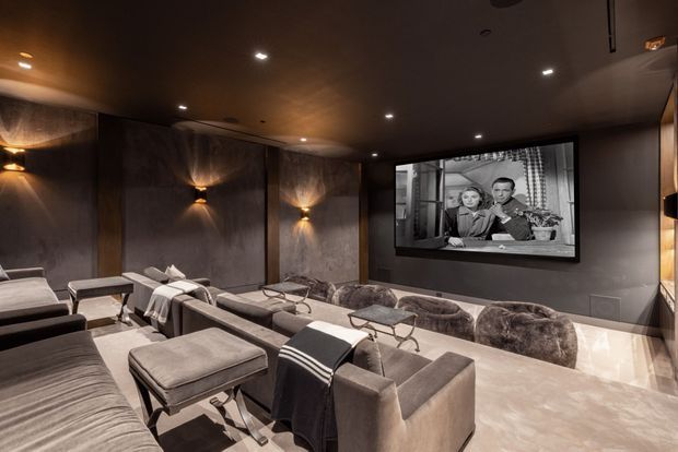 A dark, luxurious home cinema with tiered rows of plush upholstered seating, ottomans, side tables, recessed ceiling lights, wall sconces, dark textured wall panels, and a large wall-mounted screen displaying a film. The room has a warm, intimate atmosphere with polished light stone flooring and dark built-in detailing.