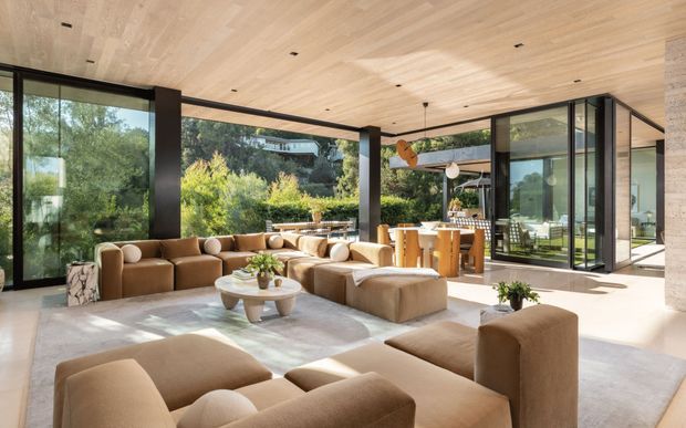 A spacious, open-plan contemporary living room with expansive floor-to-ceiling glazing, a warm pale timber parquet ceiling, dark structural columns and frames, and a large tan sectional sofa arranged around low coffee tables. The room opens visually toward a dining and kitchen area, with landscaped greenery visible beyond the glass. Natural light, neutral furnishings, and strong indoor-outdoor connection create a refined, airy atmosphere.